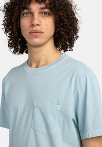 Světle modré bavlněné tričko s kulatým výstřihem a krátkými rukávy, s jednou kapsou na levé straně hrudi a hladkou, měkkou texturou.