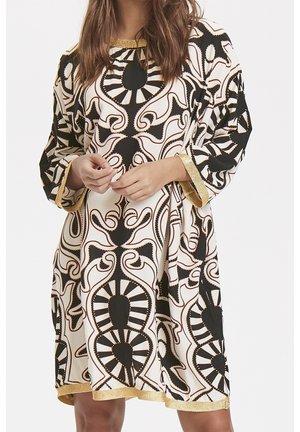 Mujer con un vestido suelto con estampado en blanco y negro y ribete dorado en las mangas y el dobladillo, con las manos entrelazadas a la altura de la cintura.