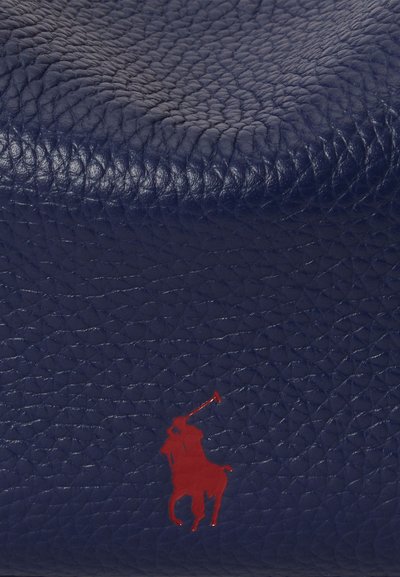 Temno modra usnjena površina z teksturiranim zaključkom, z izrazito rdečo logotipom igralca polo. Material se zdi trpežen in visokokakovosten.