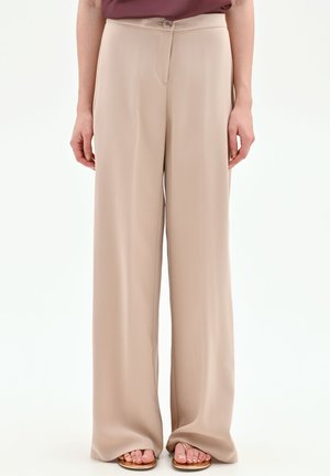 Femme portant un pantalon large beige et des sandales marron, montrée de la taille aux pieds sur un fond uni.