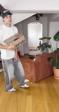 Cutie de carton cu logo-ul cerc roșu în mâini, blugi gri uzati, cămașă albă, adidași argintii, canapea portocalie din catifea, plantă verde în ghiveci.