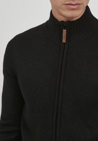 Svart zip-up genser med ribbet høy krage. Har en kontrasterende skinnlapp på glidelåsen. Laget av et teksturert strikket stoff.