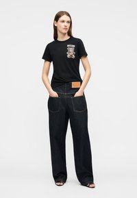 Femme portant un t-shirt noir Moschino avec un motif d'ours en peluche et un jean taille haute à jambes larges de couleur foncée avec de grandes poches arrière.