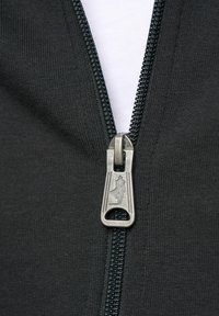 Sorte glidelåsjakke med et sølvfarget metallglidelåsholder, med logo. Stoffet har en glatt tekstur, med et kontrasterende hvitt stoff under.