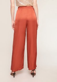 Kvinna i högmidjade, vida rostfärgade satintrosor och svarta klacksandaler, stående mot en ljus, enfärgad bakgrund.