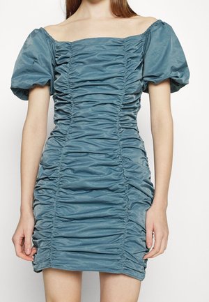 Femme portant une robe turquoise drapée, épaules dénudées, manches courtes bouffantes, silhouette cintrée et tissu texturé.