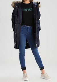 Námořnický prošívaný parka s límcem z umělé kožešiny, černé tričko s grafikou, modré skinny džíny a světlé tenisky. Obsahuje zipové a patkové uzávěry.