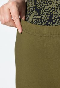 Olivově zelené legíny s širokým páskem, vyrobené z měkké, elastické tkaniny, s hladkými švy a minimálními detaily textury.