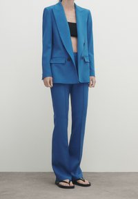 Blue tailored blazer with broad lapels and two front pockets, paired with matching flared trousers and black sandals. Smooth texture.