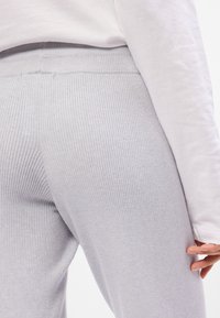 Calças em cinza claro com textura canelada, cintura ajustada, tecido texturizado e um ajuste justo, parcialmente visíveis ao lado de uma blusa branca de mangas longas.