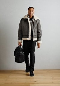 Γκρι τζάκετ από shearling με άσπρη γούνα στον γιακά, μαύρες λεπτομέρειες από φερμουάρ, συνδυασμένο με μαύρα παντελόνια και μια μαύρη τσάντα duffle. Φόντο από ξύλινο δάπεδο.