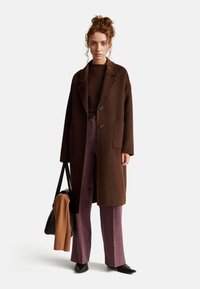 Brązowy wełniany płaszcz z dużymi kieszeniami naszytymi i kołnierzykiem z klapami, zestawiony z spodniami w szaro-fioletową kratę i brązową bluzką. Czarna torebka.