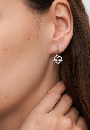 Sølv hoop-ørering med et sammenflettet hjerte-design med klare accenter, der hænger fra en enkel låselukning.