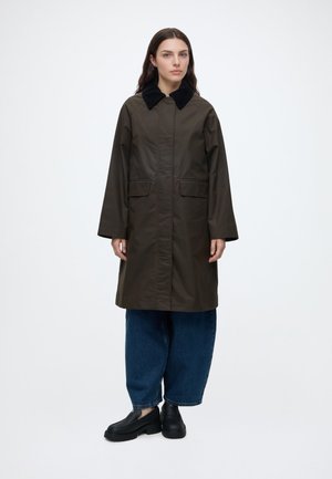 Femme portant un long manteau olive foncé avec un col noir, un jean bleu ample et des chaussures noires, debout devant un fond blanc uni.
