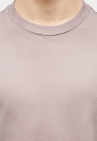 Κοντινό πλάνο του λαιμού και του άνω στήθους ενός ατόμου που φοράει ένα βαμβακερό μπλουζάκι με στρογγυλή λαιμόκοψη ανοιχτό μωβ.