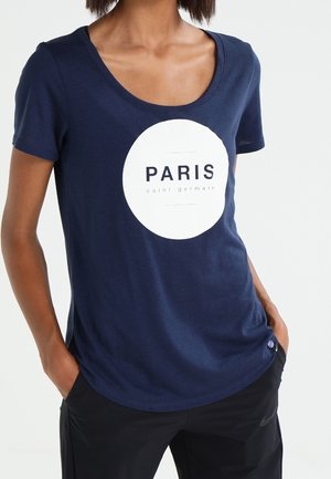 Femme portant un t-shirt bleu marine avec un cercle blanc et les mots "PARIS saint-germain" imprimés à l'avant, mains dans les poches d'un pantalon noir.