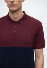 Maroon and navy polo with a textured fabric, three brown buttons, and a small blue emblem on the chest. Short sleeves and a classic collar.