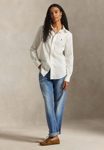 Femme aux longs cheveux bouclés portant une chemise blanche boutonnée, un jean bleu retroussé et des mocassins marron, debout devant un fond uni.