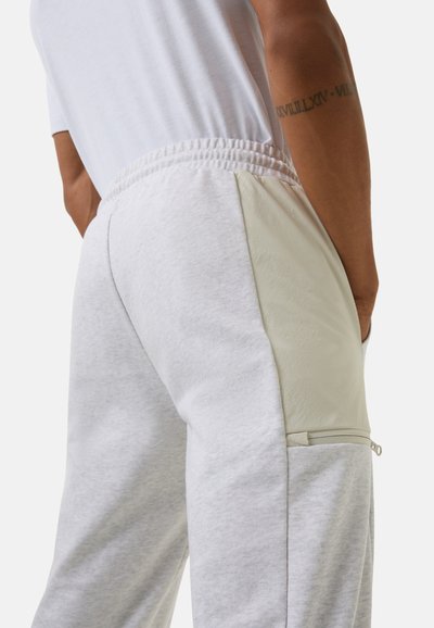 Hellgraue Sweatpants mit strukturierter Oberfläche, elastischem Bund und einer Reißverschlusstasche auf einem glatten cremefarbenen Einsatz. Kontrastnahtdetail.