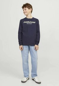 Cămașă bleumarin cu mâneci lungi și logo alb, blugi deschiși la culoare și adidași albi. Cămașa are guler rotund și croială normală.