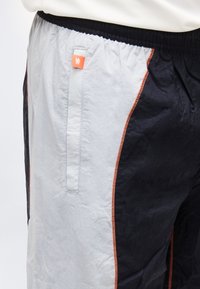 Κοντινό πλάνο μαύρου και ανοιχτόγκρι παντελονιού με πορτοκαλί ραφές και πορτοκαλί ετικέτα κοντά στην πλαϊνή τσέπη, φορεμένο με λευκό τοπ.