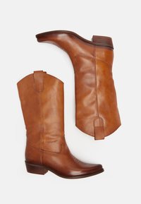 Botas de cowboy de cuero marrón con puntas afiladas, detalles de costura en los tubos y tacón apilado. Cuenta con lengüetas para facilitar su uso.