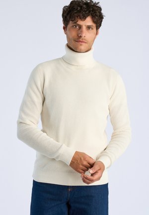 Mand med krøllet hår iført en hvid strikket polotrøje og blå jeans, justerer sin ærme mod en ensfarvet lys baggrund.