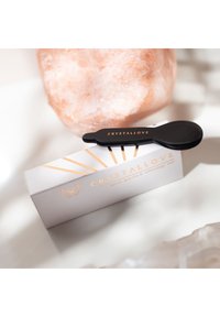 Czarny narzędzie gua sha o zaokrąglonym kształcie spoczywa na jasnoszarej pudełku z złotymi akcentami i napisem "CRYSTALLOVE." Tło składa się z różowego kryształu solnego.