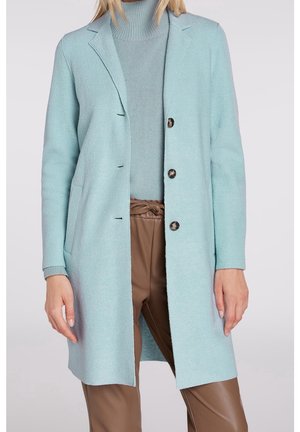 Mujer con abrigo largo azul claro sobre suéter de cuello alto a juego y pantalones de cuero marrón de talle alto con cinturón atado.