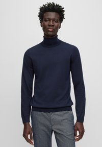 Marinblått polokrage-tröja i fint stickat material, med figursydd silhuett och långa ärmar, kombinerad med strukturerade gråbyxor.