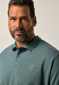 Zelená polo trička s límečkem, třemi knoflíky a jemným logem na hrudi. Hladký textilní materiál s přizpůsobeným střihem.