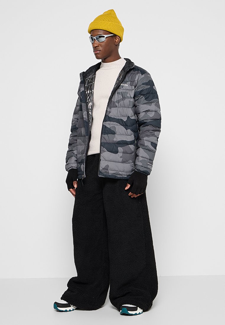 Chaqueta acolchada en gris camuflaje con un forro interior negro brillante. Lleva puesta sobre un suéter de punto crema y combinada con pantalones de forro polar negros y zapatillas.