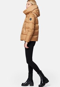 Brązowa kurtka puchowa z wysokim kołnierzem, błyszczącym wykończeniem, bocznymi kieszeniami i okrągłą czarną łatą na rękawie; zestawiona z czarnymi wąskimi dżinsami i masywnymi czarnymi butami.