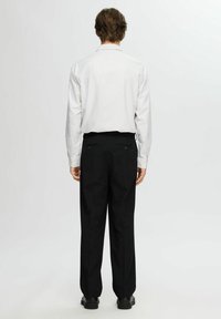 Man som står med ryggen mot kameran, iklädd en vit randig långärmad skjorta, svarta kostymbyxor och svarta skor mot en neutral bakgrund.