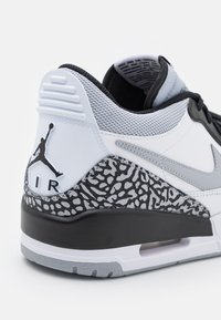 Bílý sneaker s černými a šedými akcenty. Má texturovaný černobílý vzor na patě, šedou síťovinovou svršek a viditelnou vzduchovou jednotku.