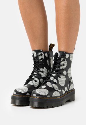Bottes en cuir noir à la cheville avec une semelle épaisse, présentant un motif de cercles et de rayures blanches, ainsi que des lacets noirs. Coutures jaunes en accent.