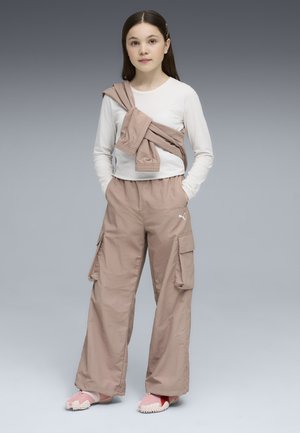 Ung pige med langt hår iført hvid langærmet skjorte, beige cargobukser, beige jakke smidt over skulderen og lyserøde sko, stående med hænderne i lommerne.