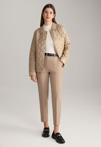 Beige doorgestikte jas met gouden drukknoopjes, gedragen over een witte geribde top en lichtbeige high-waisted broek; gecombineerd met zwarte instappers.