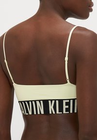 Lys gul bralette med tynne stropper og sort elastisk bånd med "CALVIN KLEIN" tekst i kremfarge. Glatt materiale, minimalistisk design.