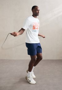 Valge Nike t-särk punase ja rohelise tekstiga, tumesinised lühikesed püksid ja valged tossud. Isik hüppab köiega heleda seina ees.