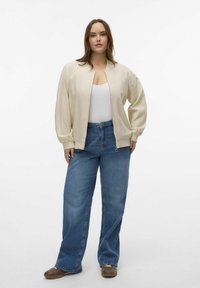 Light beige bomber jacket with a smooth texture, zip closure, and ribbed cuffs paired with mid-blue wide-leg jeans and brown shoes.