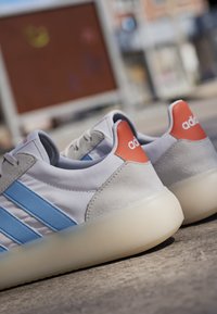 Hvide Adidas sneakers med blå striber og orange hælflapper, der hviler på en betonoverflade udendørs.