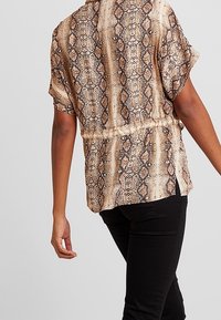 Blusa a maniche corte di colore beige chiaro con motivo a stampa serpente. Presenta una vestibilità ampia e arricciature in vita, realizzata in tessuto morbido.