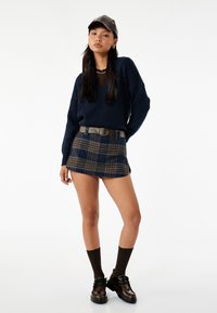 Granatowy sweter dzianinowy, mini spódniczka w kratę z niebieskimi i brązowymi akcentami, brązowy pasek, brązowe skarpetki, czarne buty na sznurówki i czapka.