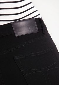 Jeans de mezclilla negra con una etiqueta de cinturón de cuero. Las características incluyen un corte ajustado, costuras sutiles y bolsillos traseros con bordes angulados.