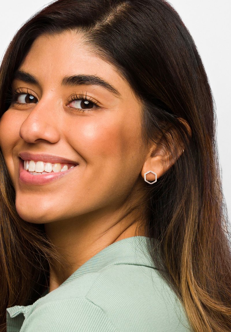 Srebrne sześciokątne kolczyki na studzie prezentowane na modelce, na jasnym tle. Modelka ma na sobie jasną zieloną koszulę z kołnierzykiem.