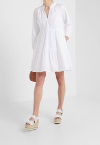 Bílá bavlněná šaty s volánovým výstřihem, dlouhými rukávy a vícevrstvou sukní. Kombinované s bílými platformovými sandály a pletenou taškou.