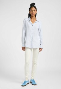 Hellblau gestreiftes Hemd mit langen Ärmeln, getragen über eng anliegenden, off-white Hosen und blauen Sneakers. Glattes Material mit legerem Schnitt.