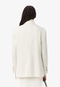 Persona con capelli intrecciati, che indossa un blazer bianco sporco e una gonna plissettata, tiene in mano una piccola borsa verde, di spalle rispetto alla telecamera.