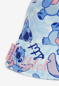 Sininen kangashattu, jossa on toistuva Stitch-kuvio Disneyn Lilo ja Stitch -elokuvasta, ja jossa on vaaleanpunaista brodeerattua tekstiä sekä -tunniste.